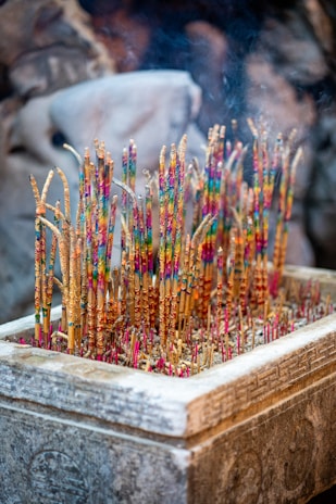A stone incense burner filled with colorful incense sticks, each adorned with vibrant patterns. Smoke subtly rises from the tips, creating a serene and mystical atmosphere. The surrounding background is softly blurred, suggesting a setting that might be spiritual or ceremonial.