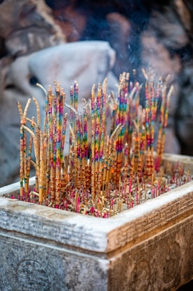 A stone incense burner filled with colorful incense sticks, each adorned with vibrant patterns. Smoke subtly rises from the tips, creating a serene and mystical atmosphere. The surrounding background is softly blurred, suggesting a setting that might be spiritual or ceremonial.