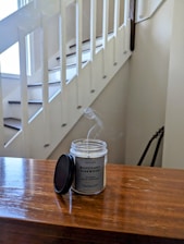 A wooden surface with a mahogany teakwood candle sitting on it, emitting a subtle wisp of smoke. The candle lid is placed beside it. The background shows a white staircase with a railing, and there appears to be a guitar leaning against the wall.