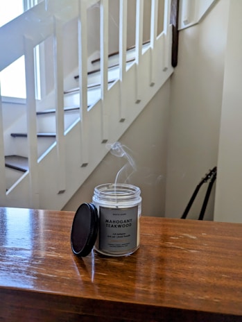A wooden surface with a mahogany teakwood candle sitting on it, emitting a subtle wisp of smoke. The candle lid is placed beside it. The background shows a white staircase with a railing, and there appears to be a guitar leaning against the wall.