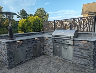 An outdoor kitchen setup featuring a stainless steel barbecue grill embedded in a stone countertop. A patio heater stands to the left, and a decorative metal screen frames the back. The area is surrounded by lush greenery with trees and bushes visible in the background.
