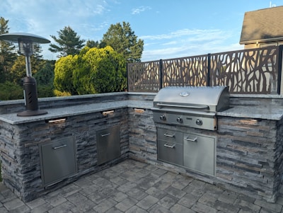 An outdoor kitchen setup featuring a stainless steel barbecue grill embedded in a stone countertop. A patio heater stands to the left, and a decorative metal screen frames the back. The area is surrounded by lush greenery with trees and bushes visible in the background.