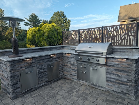 An outdoor kitchen setup featuring a stainless steel barbecue grill embedded in a stone countertop. A patio heater stands to the left, and a decorative metal screen frames the back. The area is surrounded by lush greenery with trees and bushes visible in the background.