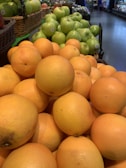 A pile of fresh grapefruits is displayed in the foreground, with green apples stacked on the table behind them. Both fruits are showcased in baskets at a grocery store setup.