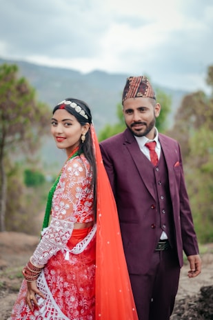 A man and woman are dressed in traditional attire, with the woman wearing a vibrant red and white embroidered dress and a red veil, and the man in a maroon suit and patterned hat. They are standing outdoors with a backdrop of blurred greenery and mountains, suggesting a serene natural setting.