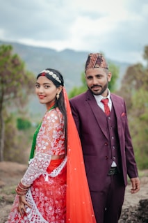 A man and woman are dressed in traditional attire, with the woman wearing a vibrant red and white embroidered dress and a red veil, and the man in a maroon suit and patterned hat. They are standing outdoors with a backdrop of blurred greenery and mountains, suggesting a serene natural setting.