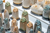 Rows of uniquely shaped stone figurines with detailed, colorful faces in display. Each figurine is detailed with engraved text, and they all have a whimsical facial expression with stylized features. The colors mainly include shades of green, grey, and orange. The figurines are arranged in a neat grid on a flat surface, and each one sits on a small individual base.
