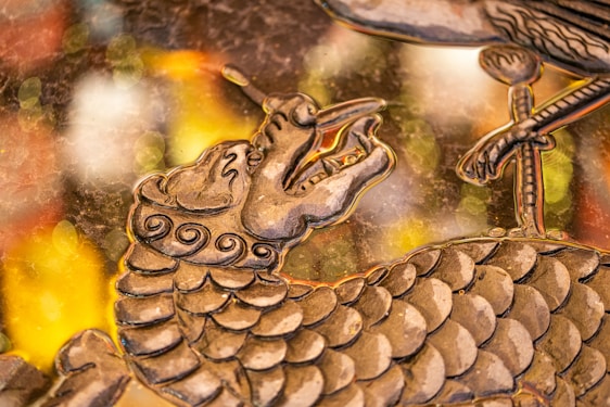 A detailed artistic depiction of a dragon's head and upper body with intricate scales and swirling patterns. The dragon has an open mouth and elongated snout. In the foreground, there is a bird's claw resting on some part of the dragon's body. The background consists of blurred, colorful bokeh lights in shades of yellow, orange, and white, giving the piece a dynamic and elegant appearance.