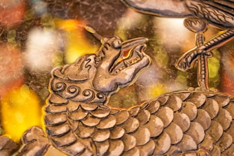 A detailed artistic depiction of a dragon's head and upper body with intricate scales and swirling patterns. The dragon has an open mouth and elongated snout. In the foreground, there is a bird's claw resting on some part of the dragon's body. The background consists of blurred, colorful bokeh lights in shades of yellow, orange, and white, giving the piece a dynamic and elegant appearance.
