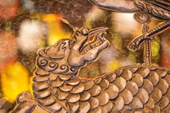 A detailed artistic depiction of a dragon's head and upper body with intricate scales and swirling patterns. The dragon has an open mouth and elongated snout. In the foreground, there is a bird's claw resting on some part of the dragon's body. The background consists of blurred, colorful bokeh lights in shades of yellow, orange, and white, giving the piece a dynamic and elegant appearance.