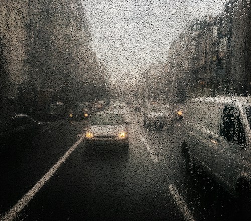 A street scene viewed through a rain-covered window, blurring the image of cars with headlights on. The overall tone is dark and moody with a focus on the abstract patterns formed by the raindrops.