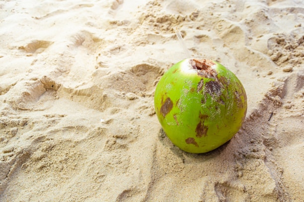 A green coconut lies on sandy beach terrain, surrounding it are ripples and footprints in the sand. The coconut has some natural brown patches and appears fresh with a small opening at the top.
