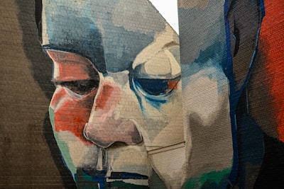 A large mural on a brick wall features an expressive and abstract portrait with bold strokes of various colors such as blue, red, beige, and white. The face, partially obscured by an adjoining wall, displays a solemn expression and is depicted with an intense focus on the eyes and nose.