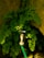 A metal water tap is attached to a green garden hose, surrounded by lush green fern leaves. The background features a textured stone wall that adds a natural, rustic element to the setting. Sunlight filters through, highlighting the leaves and the water tap.