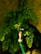 A metal water tap is attached to a green garden hose, surrounded by lush green fern leaves. The background features a textured stone wall that adds a natural, rustic element to the setting. Sunlight filters through, highlighting the leaves and the water tap.