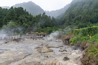 A scenic view of a geothermal area with steam rising from the ground. The landscape is lush and green with dense forested hills in the background. A wooden walkway runs across the scene, with a couple of people walking on it, observing the steamy grounds.