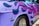 A close-up view of a vintage car with a vibrant custom paint job. The car's surface features purple and pink flame motifs. The visible chrome details include a distinctive emblem and branding on the side. The tire and rim are also partially visible.