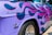 A close-up view of a vintage car with a vibrant custom paint job. The car's surface features purple and pink flame motifs. The visible chrome details include a distinctive emblem and branding on the side. The tire and rim are also partially visible.
