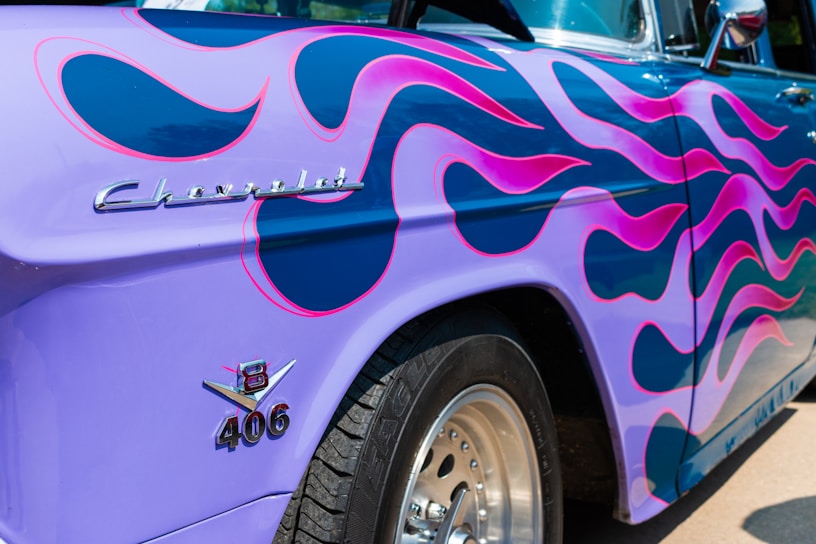 A close-up view of a vintage car with a vibrant custom paint job. The car's surface features purple and pink flame motifs. The visible chrome details include a distinctive emblem and branding on the side. The tire and rim are also partially visible.