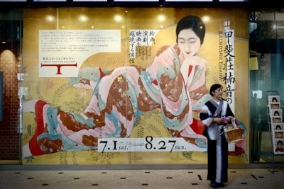 A large advertisement poster features a traditional, artistic depiction of a woman in a colorful kimono, displayed on a glass wall next to a brick wall. A real person, also dressed in a kimono, stands in front of the poster holding a basket and some papers. Japanese text is visible, including dates and references to the Tokyo Station Gallery.