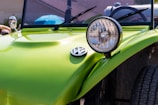 A close-up view of the front section of a lime green Volkswagen vehicle, specifically focusing on the round headlight and the Volkswagen emblem on its hood. The background shows parts of the windshield and side mirrors, with an out-of-focus blue object and a partially visible person.