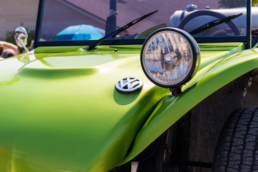 A close-up view of the front section of a lime green Volkswagen vehicle, specifically focusing on the round headlight and the Volkswagen emblem on its hood. The background shows parts of the windshield and side mirrors, with an out-of-focus blue object and a partially visible person.