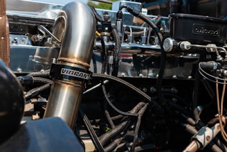 A close-up view of a car engine with visible metal pipes and various connected components. The shiny metallic surfaces reflect the surrounding environment, and a large pipe prominently displays the brand 'Vibrant'. Hoses and wires are intricately arranged around the machinery, giving a complex and industrial aesthetic.