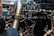 A close-up view of a car engine with visible metal pipes and various connected components. The shiny metallic surfaces reflect the surrounding environment, and a large pipe prominently displays the brand 'Vibrant'. Hoses and wires are intricately arranged around the machinery, giving a complex and industrial aesthetic.