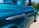 A close-up view of a vintage Chevrolet 1300 truck with a shiny, teal-blue finish. The vehicle features chrome trim details and a side mirror, set against a background of greenery and sky.