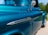 A close-up view of a vintage Chevrolet 1300 truck with a shiny, teal-blue finish. The vehicle features chrome trim details and a side mirror, set against a background of greenery and sky.
