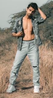 A man with a casual pose stands in a field, wearing ripped jeans and an open denim jacket, highlighting a relaxed and rugged style.