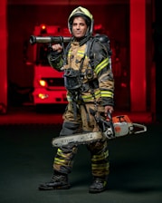 A firefighter stands confidently while wearing full protective gear, including a helmet and reflective striped uniform. He holds a chainsaw in one hand and a hose nozzle over his shoulder. A fire truck with red lights is visible in the background, creating a dramatic scene.