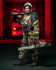 A firefighter stands confidently while wearing full protective gear, including a helmet and reflective striped uniform. He holds a chainsaw in one hand and a hose nozzle over his shoulder. A fire truck with red lights is visible in the background, creating a dramatic scene.