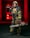 A firefighter stands confidently while wearing full protective gear, including a helmet and reflective striped uniform. He holds a chainsaw in one hand and a hose nozzle over his shoulder. A fire truck with red lights is visible in the background, creating a dramatic scene.