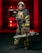 A firefighter stands confidently while wearing full protective gear, including a helmet and reflective striped uniform. He holds a chainsaw in one hand and a hose nozzle over his shoulder. A fire truck with red lights is visible in the background, creating a dramatic scene.