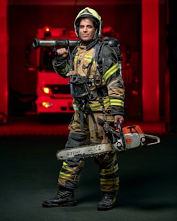 A firefighter stands confidently while wearing full protective gear, including a helmet and reflective striped uniform. He holds a chainsaw in one hand and a hose nozzle over his shoulder. A fire truck with red lights is visible in the background, creating a dramatic scene.