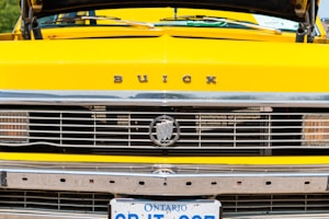 A close-up view of the front grille and license plate area of a yellow Buick car. The emblem, grille details, and bright color are prominently featured. The Ontario license plate is partially visible at the bottom.