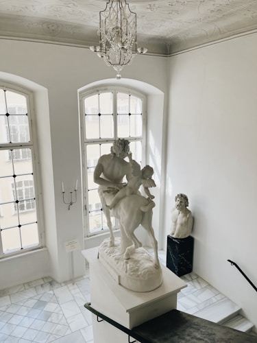 A grand staircase adorned with a large classical sculpture depicting a man and a centaur. The setting is an elegant interior with high ceilings, intricate plasterwork, and large windows that allow soft natural light to illuminate the room. A second bust sculpture is placed on a pedestal near the stair landing. A chandelier adds an element of luxury to the space.