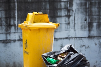 A yellow outdoor trash bin stands against a weathered, gray concrete wall. Beside it, a black garbage bag is partially open, with some green plastic and brown, decomposing leaves visible.