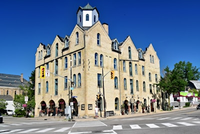 A large, historic hotel building with an off-white brick facade situated at a street corner. The architectural style features multiple peaked roofs and arched windows, with a small tower on top. The street is lined with hanging flower baskets and people walking. Various street signs and traffic lights are visible. The sky is clear and blue.