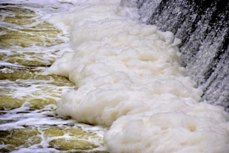 A cascading waterfall generates a thick layer of white foam as it flows over a rocky surface. The water appears turbulent, creating frothy waves and bubbles as it spreads across the scene.