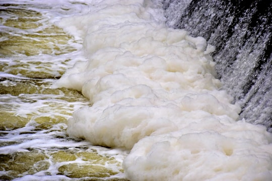 A cascading waterfall generates a thick layer of white foam as it flows over a rocky surface. The water appears turbulent, creating frothy waves and bubbles as it spreads across the scene.