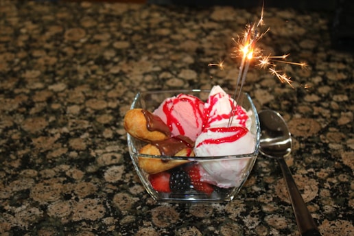 A glass bowl filled with scoops of ice cream, topped with red syrup and surrounded by sweet pastries drizzled with chocolate sauce. There is a sparkling candle inserted into the dessert, illuminating it. A spoon lies beside the bowl on a granite countertop.