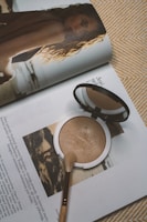 A makeup compact and brush rest on the pages of an open magazine. The magazine features an image of a woman with wavy hair looking back over her shoulder at the camera. Text is visible on the pages, and the overall setting creates a sense of everyday beauty and style.