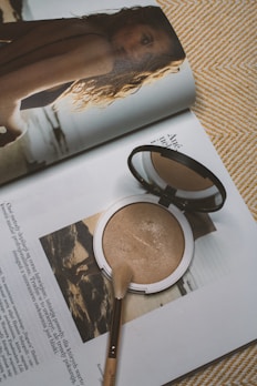 A makeup compact and brush rest on the pages of an open magazine. The magazine features an image of a woman with wavy hair looking back over her shoulder at the camera. Text is visible on the pages, and the overall setting creates a sense of everyday beauty and style.