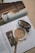 A makeup compact and brush rest on the pages of an open magazine. The magazine features an image of a woman with wavy hair looking back over her shoulder at the camera. Text is visible on the pages, and the overall setting creates a sense of everyday beauty and style.