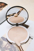 An open compact powder with a delicate brush, set against a soft beige fabric background.