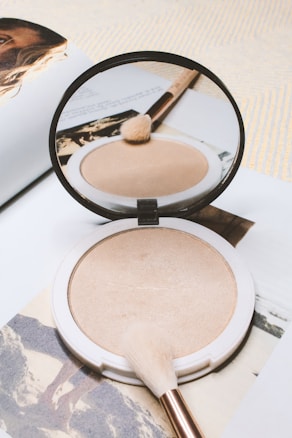 A round compact of beige powder makeup sits open on a page of an open magazine. A soft makeup brush with a light brown handle rests on the powder and is reflected in the compact's mirror.
