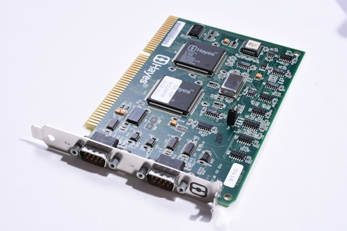 A green circuit board with multiple integrated circuits and electronic components, including resistors, capacitors, and connectors. The board features a series of gold-plated connectors on one edge and various chips labeled with 'Hayes,' indicating it is likely a computer hardware component such as a modem. The surface is populated with detailed white printed traces and black connectors for interfacing.