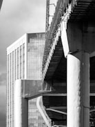 A black and white photograph captures a modern urban scene with a prominent high-rise building featuring numerous windows and reflective glass. In the foreground, there is a large concrete structure under construction, with visible beams and scaffolding. The contrast between the finished skyscraper and the developing construction creates a dynamic architectural composition.